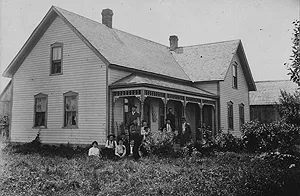 A family poses outside a two-story wooden farmhouse with a decorative front porch.