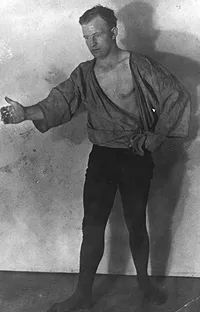 A man wearing a loose canvas jacket open at the chest and dark trousers, posing with one arm extended forward against a plain backdrop.