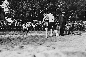 Two men wearing wrestling jackets grapple in an outdoor ring while a referee in a suit looks on, surrounded by a large crowd of spectators.