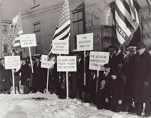 A crowd gathered in the snow outside a stone building, holding American flags and signs with slogans such as 'NO RED FLAG FOR US' and 'WE WANT WORK, PEACE, PLENTY NOT AGITATORS'.