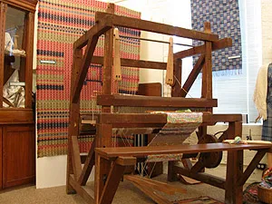 A large wooden floor loom with an attached bench and partial weaving on display in front of a wall-mounted red and blue patterned blanket.
