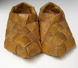 A pair of slip-on shoes crafted from wide, woven strips of light brown birch bark in a diagonal basketweave pattern.