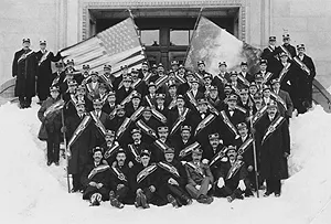 A large group of men in ceremonial sashes and caps poses on snowy steps beneath two large flags.