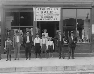 A group of men and boys stands outside a shop under a banner reading 'GRAND OPENING SALE CLOTHING, MENS FURNISHINGS & SHOES'.