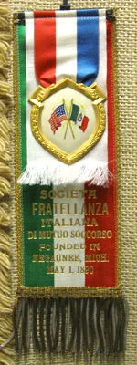 A fabric ribbon badge trimmed in gold fringe with green, white, and red stripes, featuring a central shield emblem with crossed American and Italian flags above gold text reading 'Founded in Negaunee, Mich. May 1, 1890'.