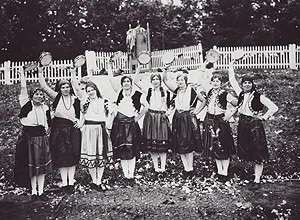 Eight women in traditional embroidered folk clothing pose in a row outdoors, each holding a tambourine overhead in front of a white picket fence.
