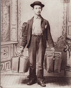 A young man in a bowler hat and jacket carrying a pack on his back and two large sample bags.
