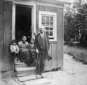 An elderly man holding a cane stands outside a simple wooden cabin beside an elderly woman and two young children seated on the porch steps.
