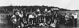 A large group of men holding brass instruments gathered in an open field near a farm building.