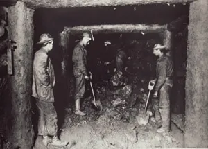 Miners with shovels working inside an underground mine shaft framed by heavy vertical timber posts and overhead support beams.