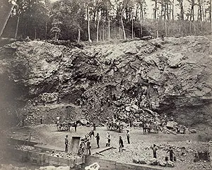 Miners and horse-drawn wagons working at the base of a tall, rocky cliff face in an open-pit iron mine.