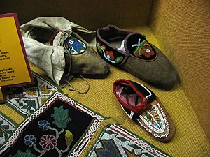 Three handcrafted leather moccasins featuring intricate floral beadwork, displayed alongside patterned beaded textiles in a museum case.