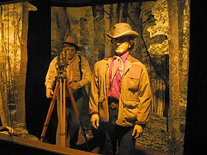 A museum diorama of two surveyors in hats with a tripod transit in a forest setting.