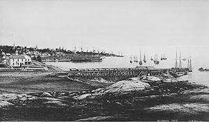 A wooden ore dock extends into a harbor filled with sailing ships along a rocky shoreline.