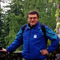 Arnold Alanen wearing glasses and a blue jacket outdoors against a backdrop of green trees.