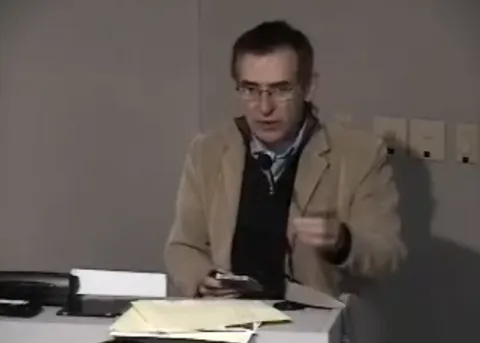 A man in glasses and a tan jacket gestures while speaking behind a lectern with papers.